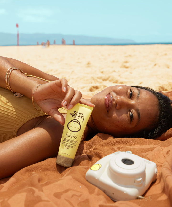 Woman lying on the beach, smiling and holding a tube of Original SPF 50 Sunscreen Face Lotion by Sun Bum.