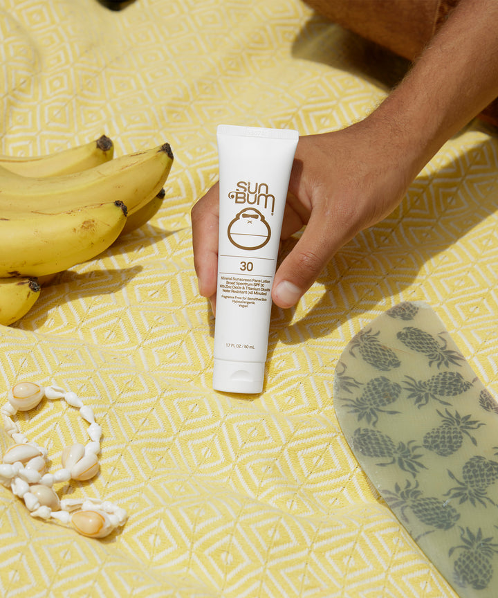 A hand holding a tube of Mineral SPF 30 Sunscreen Face Lotion by Sun Bum on a yellow patterned background next to bananas and shell accessories.