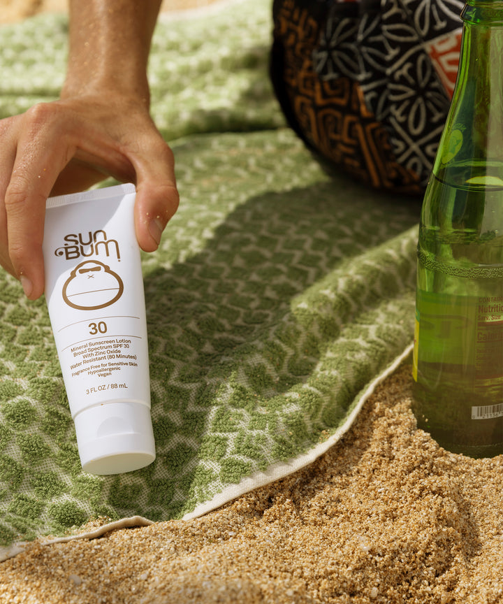 Mineral SPF 30 Sunscreen Lotion by Sun Bum on a green beach towel with a hand holding the bottle and a drink nearby.