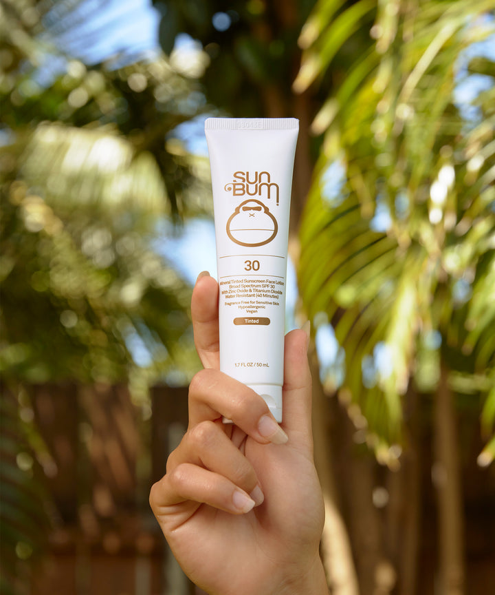 Hand holding a tube of Mineral SPF 30 Tinted Sunscreen Face Lotion with tropical palm trees in the background, Sun Bum.