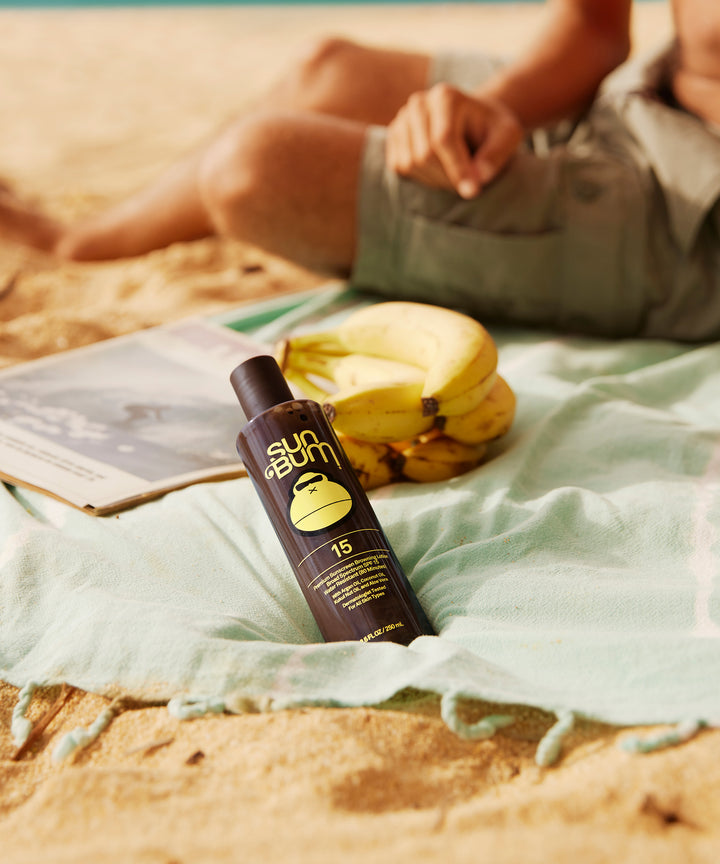 SPF 15 Sunscreen Browning Lotion by Sun Bum on a beach towel next to a bunch of bananas with a relaxed person in the background, Sun Bum