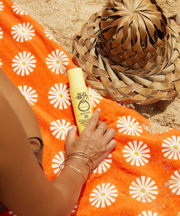 SPF 30 Sunscreen Oil by Sun Bum on an orange towel with white floral patterns, next to a woven hat, perfect for hydrating skin and protecting against UV rays.