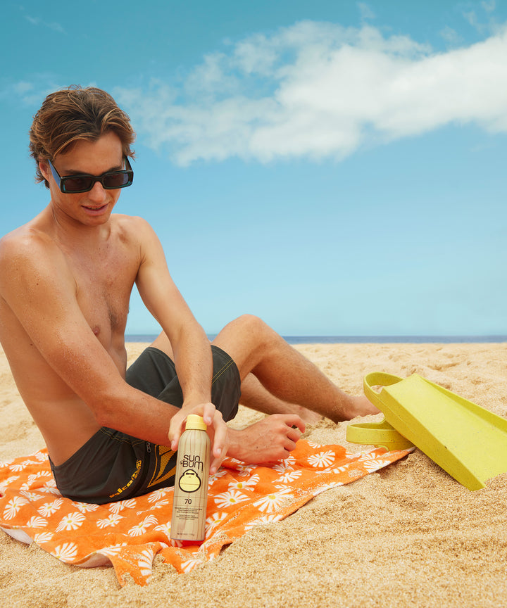 Shirtless man applying sun protection spray on the beach, Sun Bum