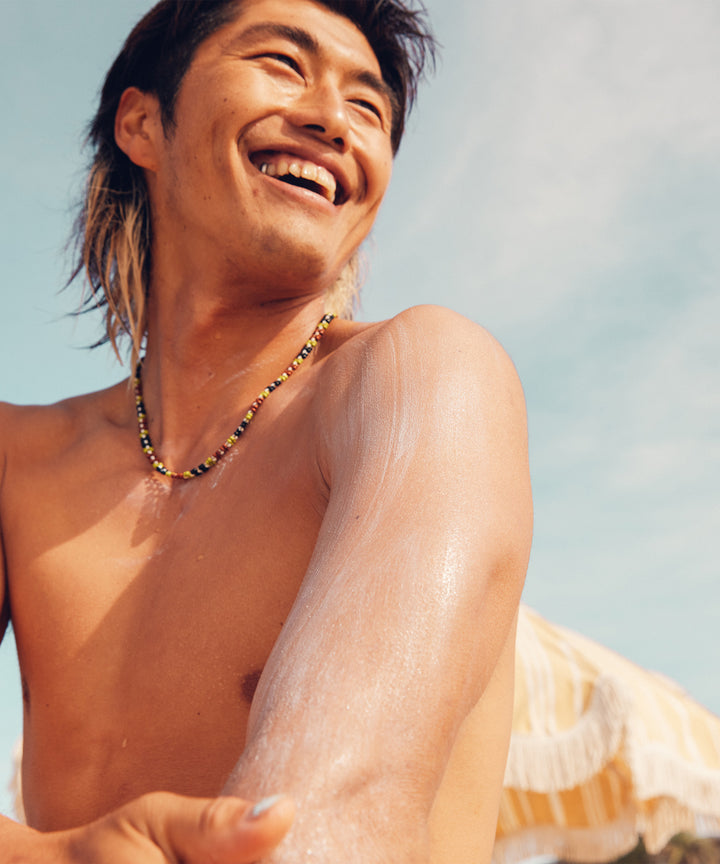 Man smiling while applying Original SPF 30 Sunscreen Lotion on his arm at the beach, Sun Bum.