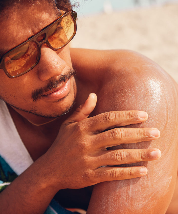 Man applying Original SPF 50 Sunscreen Lotion on his shoulder at the beach, promoting sun protection, by Sun Bum.