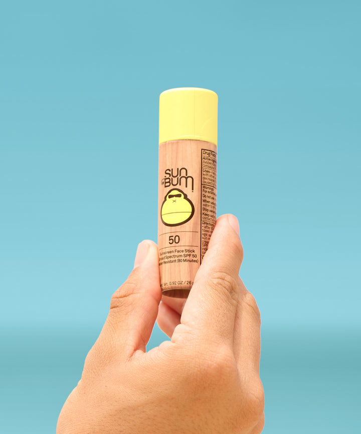 Hand holding a Paper Tube SPF 50 Sunscreen Face Stick with yellow cap on a blurred blue sky background, Sun Bum