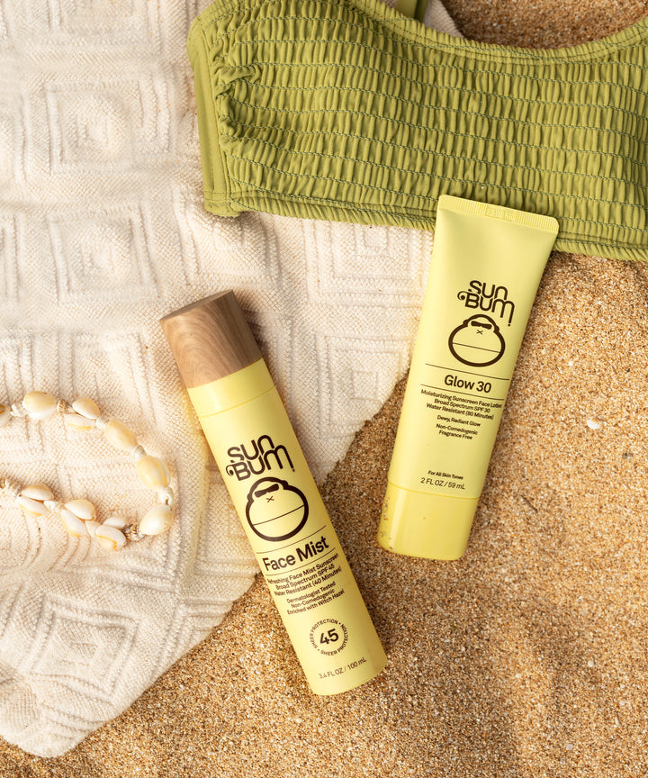 Sun Bum Original Glow SPF 30 Sunscreen Face Lotion and Face Mist on sandy beach with green top and seashell necklace.