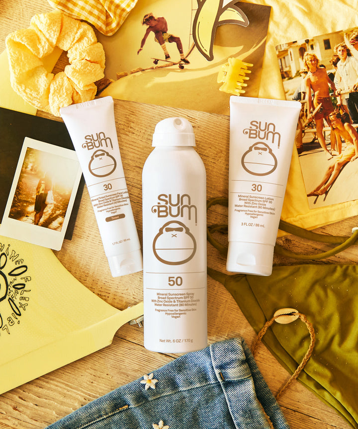 Mineral SPF 50 Sunscreen Spray by Sun Bum surrounded by summery items, showcasing its zinc-based, fragrance-free formula in a non-aerosol compression container without chemical propellants. Sun Bum