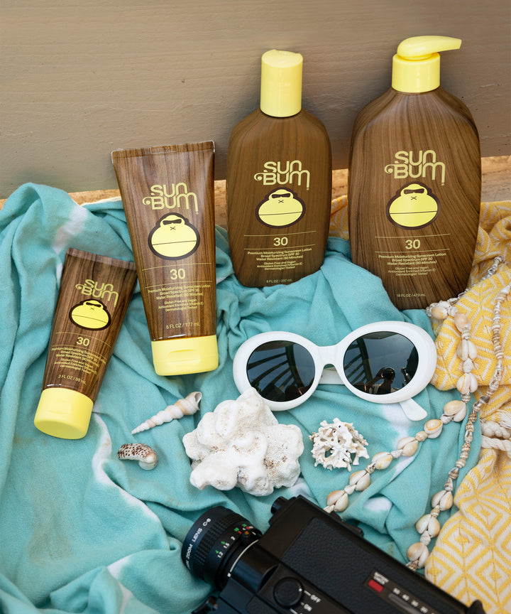 A collection of Sun Bum Original SPF 30 Sunscreen Lotion products displayed on a teal fabric along with seashells, white sunglasses, and a vintage camera. Sun Bum.