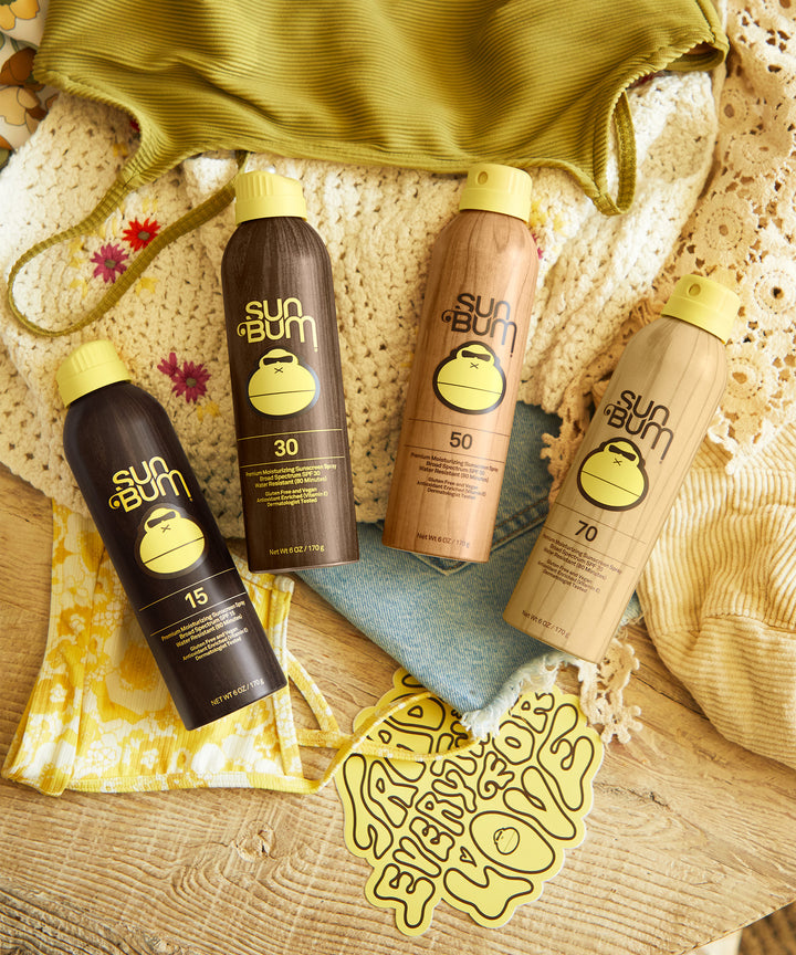 A variety of Sun Bum sunscreen spray bottles, including Original SPF 30 Sunscreen Spray, displayed on a wooden table with beach essentials like a green bag, crochet items, and a scarf. Sun Bum,