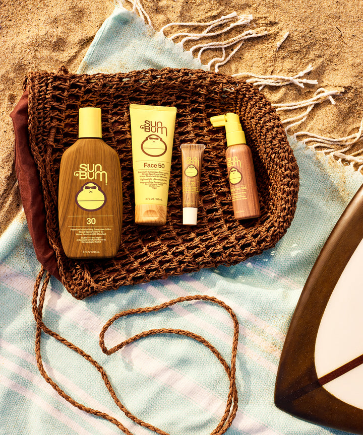 Woven beach bag containing Sun Bum sunscreen products on sand, including SPF 30 lotion and lip balm, next to blue towel and sunglasses. Perfect beach day essentials for sun protection. Sun Bum