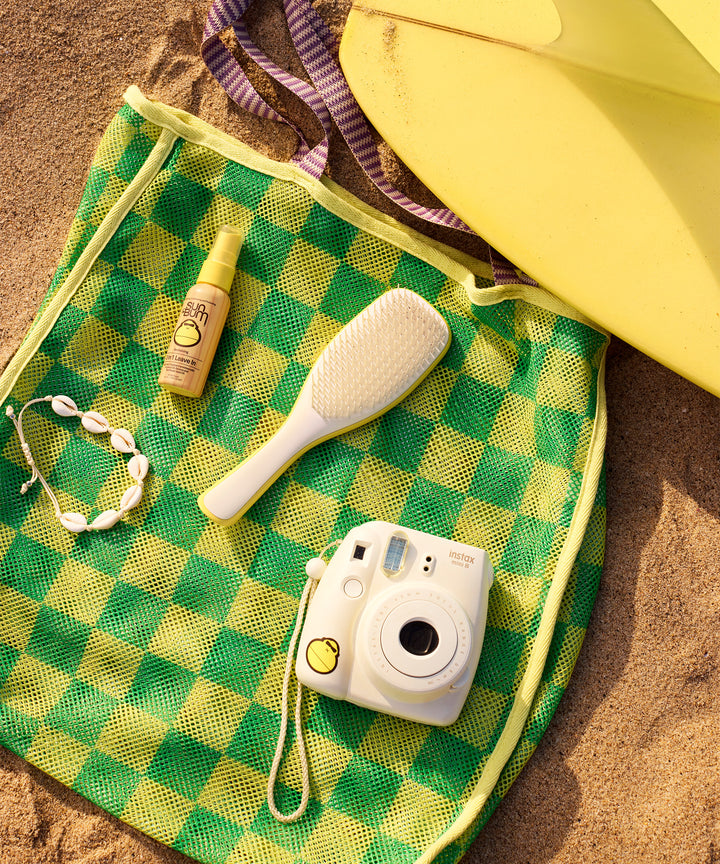 Beach essentials on green checkered bag: yellow surfboard, white detangling brush, leave-in conditioner, instant camera, and shell necklace. Perfect for a day of sun and surf with Sun Bum products.