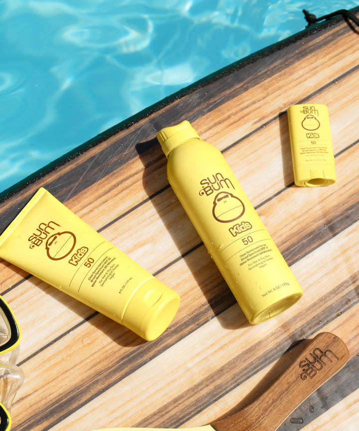 Yellow Sun Bum Kids SPF 50 Clear Sunscreen products are placed on a wooden surfboard, near a pool. Sun Bum,